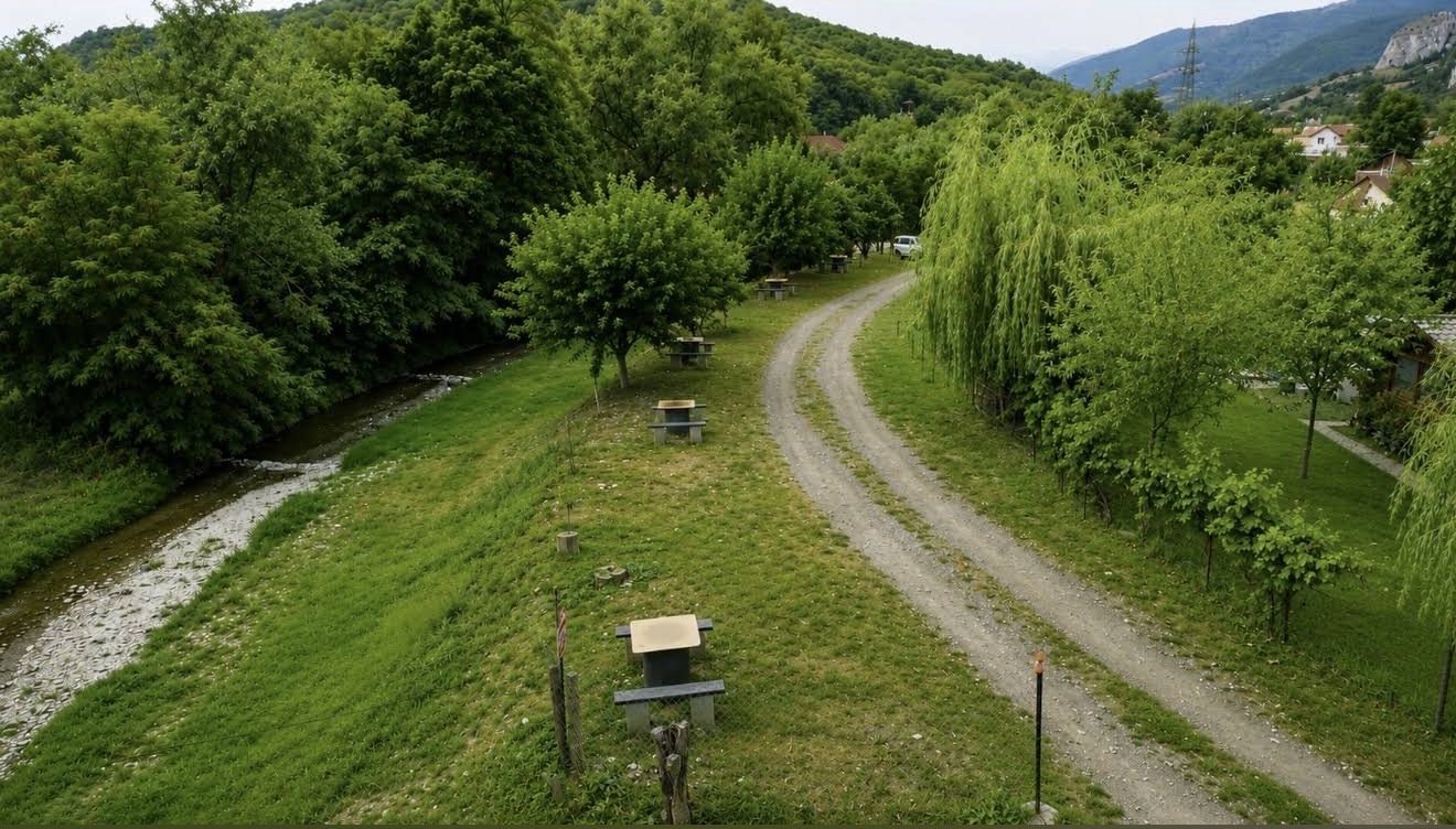 Ein Schotterweg zwischen Bach und Bäumen, mit Picknicktischen auf der Wiese